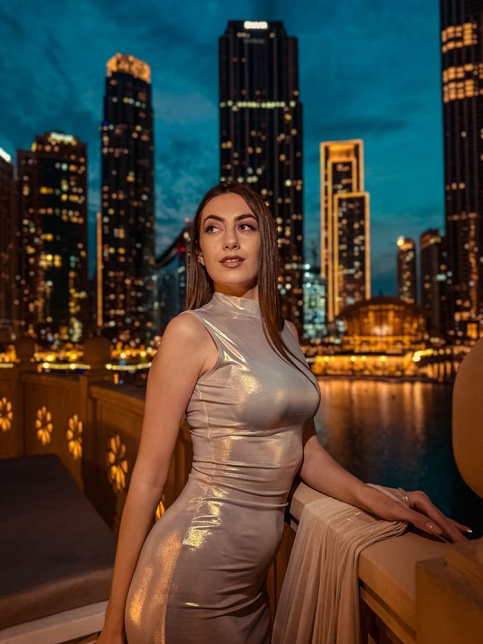 Woman in shimmering dress poses on Dubai balcony with city skyline at night.